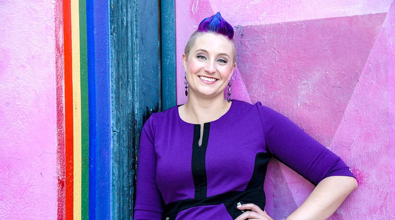 A photo of Dr. Liz Powell, sexy psychologist, standing in front of a purple wall. 