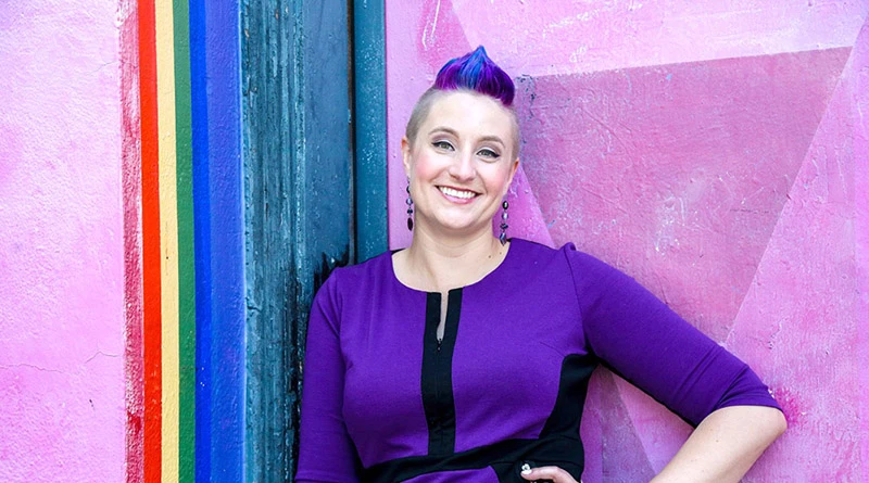 A photo of Dr. Liz Powell, sexy psychologist, standing in front of a purple wall. 