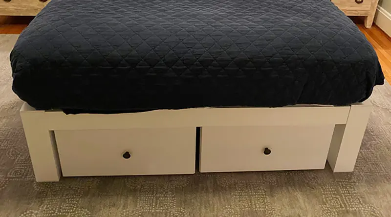 Two twelve inch high white wooden drawers underneath a white queen size metal Platform Bed with a dark blue comforter.