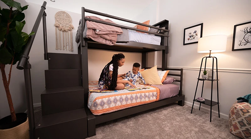 A young mother and her son reading a book on the bottom bunk of a charcoal Twin XL Over Queen Adult Bunk Bed with a staircase in their pajamas before bed.