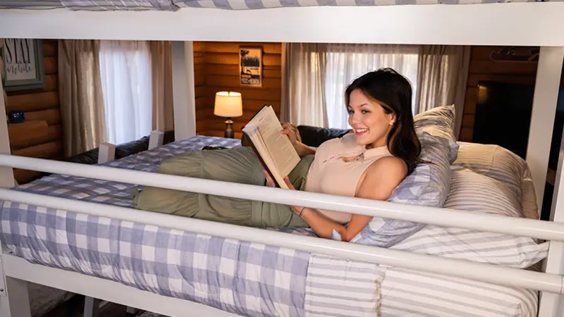 A young woman reading a book while sitting up on the middle bunk of a white queen size Triple Bunk Bed for adults.