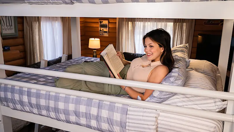 A young woman reading a book while sitting up on the middle bunk of a white queen size Triple Bunk Bed for adults.