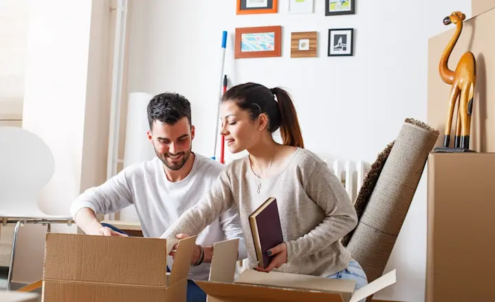 A smiling couple sits on the floor amidst cardboard boxes in a living room. They are unpacking items, including books and decor. The room features framed art on the wall and a wooden giraffe statue.