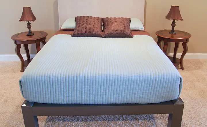 A neatly made bed with a light blue quilt and two brown pillows, flanked by wooden side tables. Each table has an ornate lamp with a brown shade, resting on a beige carpet in a room with light-colored walls.