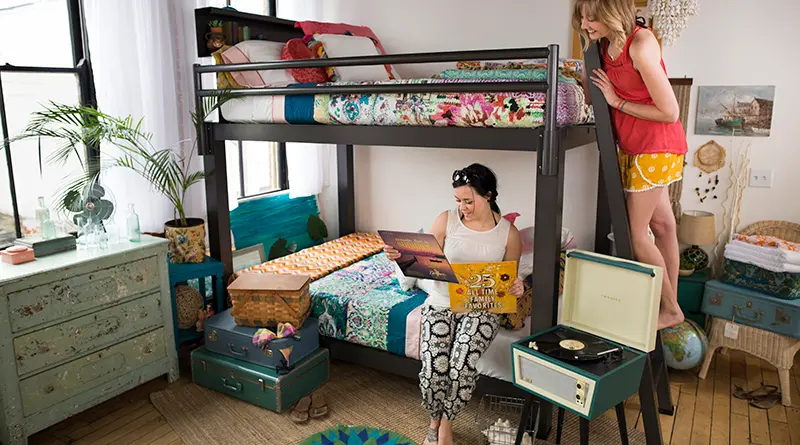 A charcoal Queen Adult Bunk Bed in the bedroom of two young adult women. One of them is sitting on the bottom bunk looking at the cover of a vinyl record and the other is standing on the ladder next to her looking over her shoulder. Seen from a slight angle toward the lower right-hand corner of the bed.