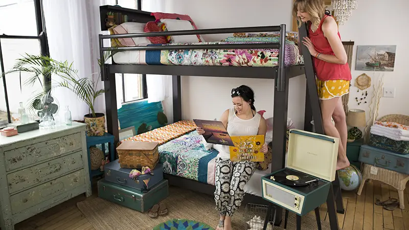 A charcoal Queen Adult Bunk Bed in the bedroom of two young adult women. One of them is sitting on the bottom bunk looking at the cover of a vinyl record and the other is standing on the ladder next to her looking over her shoulder. Seen from a slight angle toward the lower right-hand corner of the bed.