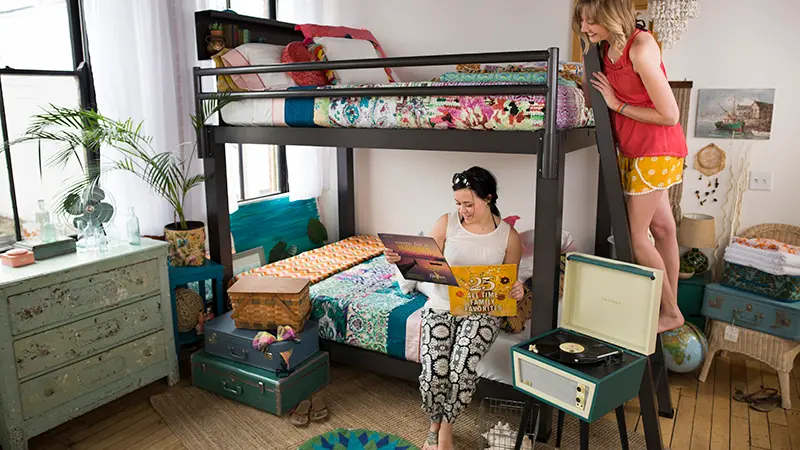 A charcoal Queen Adult Bunk Bed in the bedroom of two young adult women. One of them is sitting on the bottom bunk looking at the cover of a vinyl record and the other is standing on the ladder next to her looking over her shoulder. Seen from a slight angle toward the lower right-hand corner of the bed.