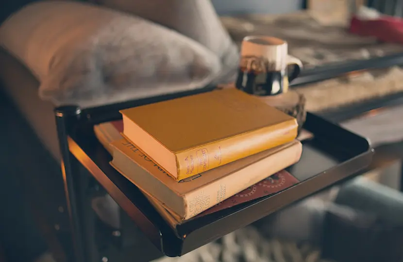 A stack of books rests on a small black tray, with a white and brown mug placed on top. The background features a cozy setting with a sofa and plush blanket, suggesting a comfortable reading nook.