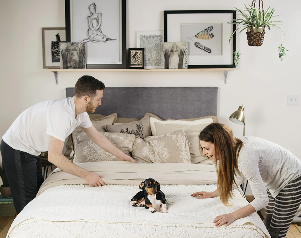 A young couple bent over each side of their white queen size metal Platform Bed with a blueish gray headboard smoothing out the blankets as they finish making the bed while they're small black dog rolls on the bed.