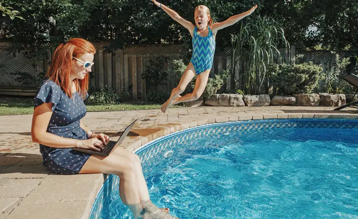 A mom sits at the ledge of a pool working on her laptop while her daughter jumps in