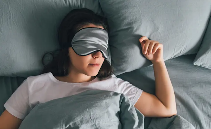 Young woman lying on a bed with blue-grey bedding wearing a matching sleep mask, seen from above at a high angle.