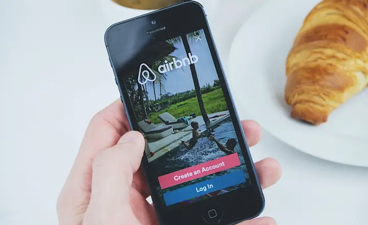 A hand holds a smartphone displaying the Airbnb app login screen. In the background, there's a cup of coffee and a croissant on a white plate, placed on a white table.