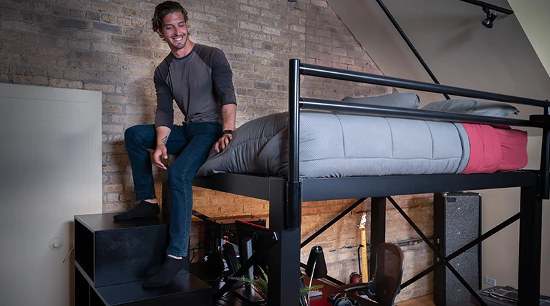 A young man sitting on the edge of his black Queen Adult Loft Bed with his feet resting on the matching staircase