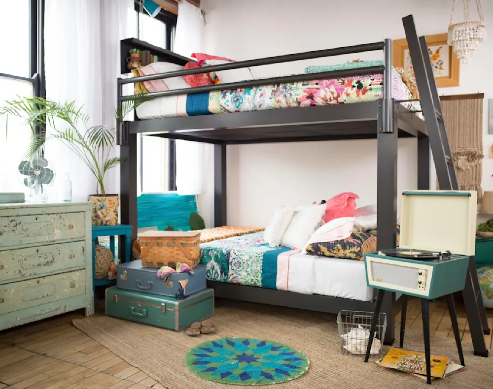 A cozy, eclectic bedroom with a metal bunk bed adorned with colorful bedding. There's a vintage dresser, a record player on a stand, and two teal suitcases. The room is decorated with plants, art, and a globe, creating a vibrant and inviting atmosphere.