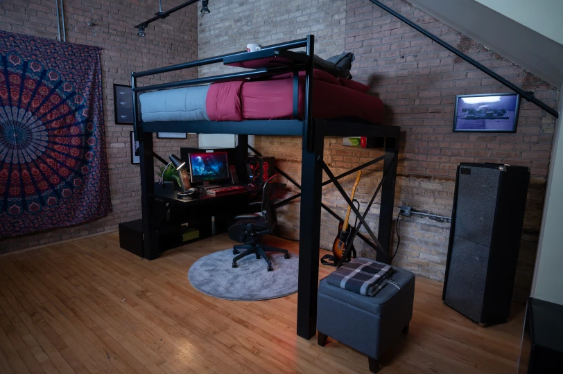 A black Loft Bed with a gaming desk below it, in a dark room with tapestry on the wall and guitar nearby.