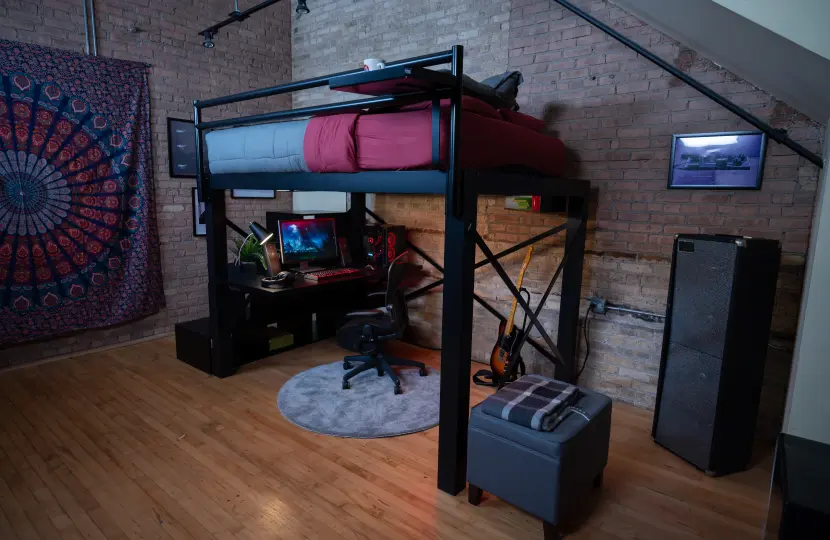 A black Loft Bed with a gaming desk below it, in a dark room with tapestry on the wall and guitar nearby.