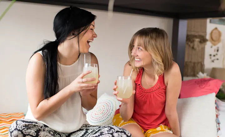 Two women sitting on a colorful couch, laughing and holding glasses of lemonade. One wears a white top and patterned pants, the other a red top and yellow shorts. The setting is relaxed and cozy.
