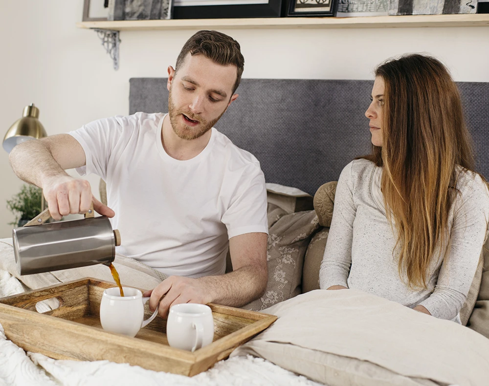 A young couple sitting up in their white queen size metal platform bed with a blueish gray headboard. They have a small wooden tray on their laps and the male is pouring coffee from a pot into a white mug on the tray.
