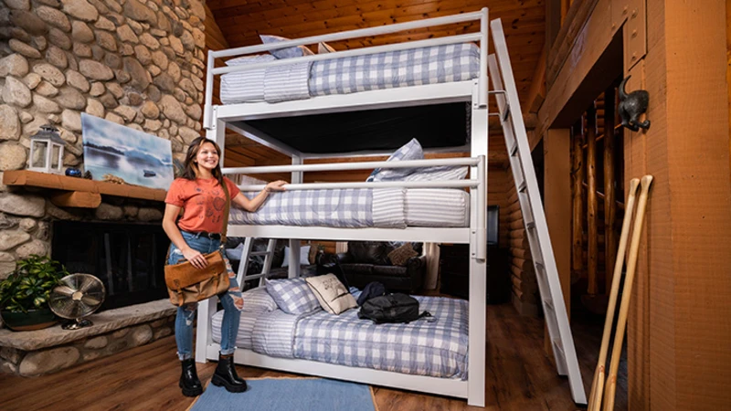 Young woman standing next to a white Triple Bunk Bed for adults with blue and white bedding inside of an upscale, rustic cabin with her left arm on the guard rail of the middle bunk.