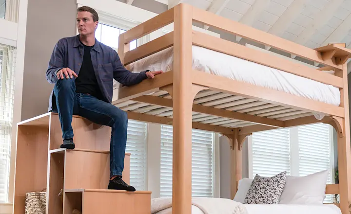 A man in casual clothes sits on wooden stairs leading to a lofted bed in a bright, modern bedroom with large windows and a bed underneath the loft.