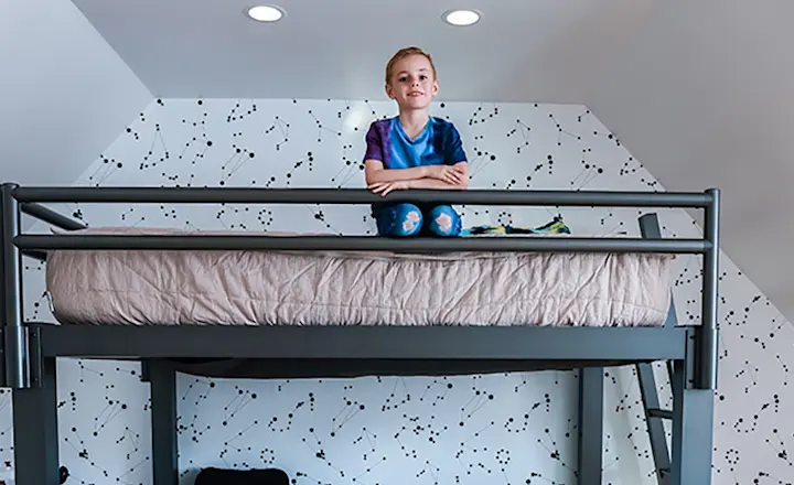 A young blonde boy sits on the top bunk of a charcoal Queen Over Queen Adult Bunk Bed and smiles down at the camera facing the bed from the side.