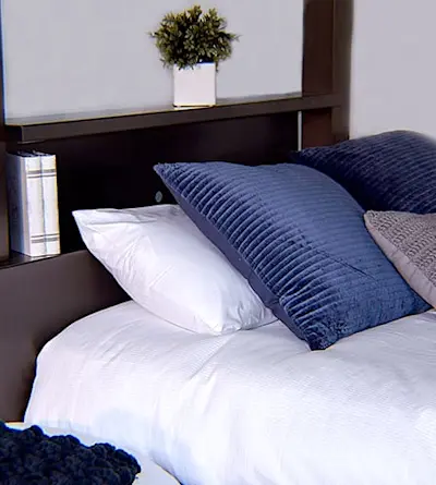 A neatly made bed with white sheets and a mix of blue and gray pillows. A small plant and a book are placed on a shelf attached to the dark wood headboard.