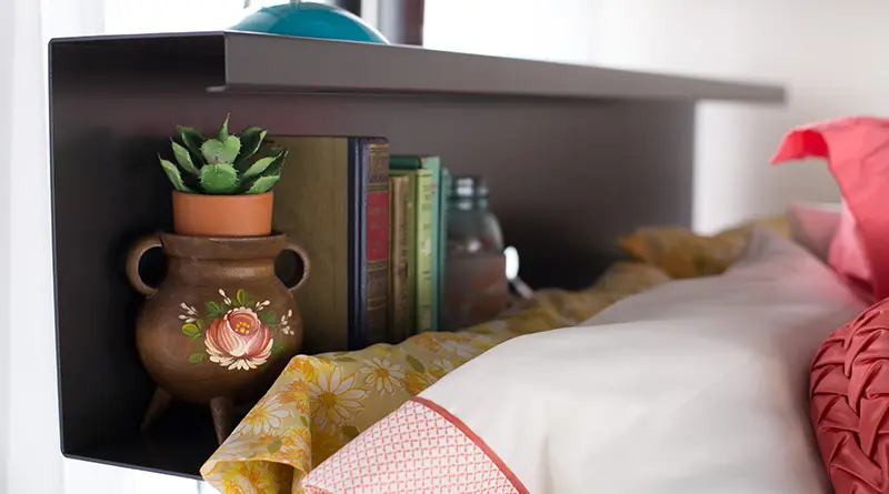Charcoal bookshelf headboard on a charcoal metal bed frame. Has some books and a small potted plant on the shelf and you can see the bottom of a blue desk lamp on the top of the bookshelf.