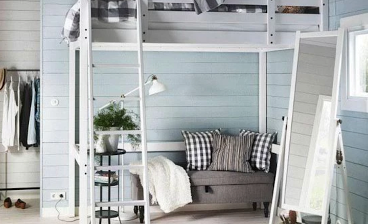 A cozy loft bedroom with a gray sofa beneath a white loft bed. The room features light blue paneled walls, a floor mirror, and a soft gray rug. A small side table with a plant and a lamp adds a touch of greenery. A clothing rack is seen in the corner.