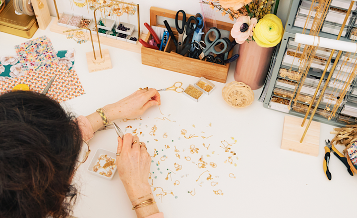 A woman sitting at her brightly lit and well-stocked craft table, making beaded jewelry.