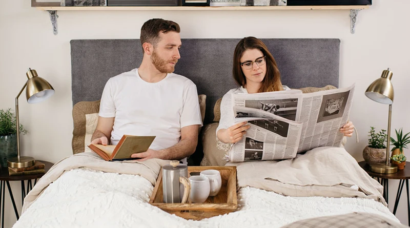 A young adult couple sitting in their white queen size Platform Bed and sitting up against the headboard reading.