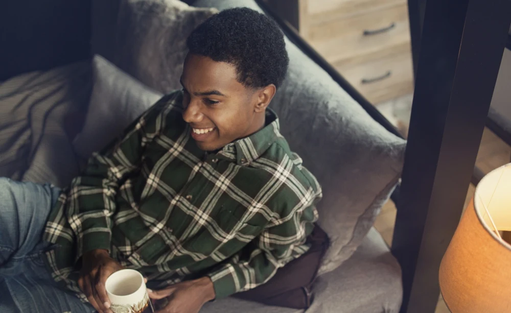 A man sits on the bottom bunk of an Adult Bunk Bed smiling and enjoying a cup of coffee.