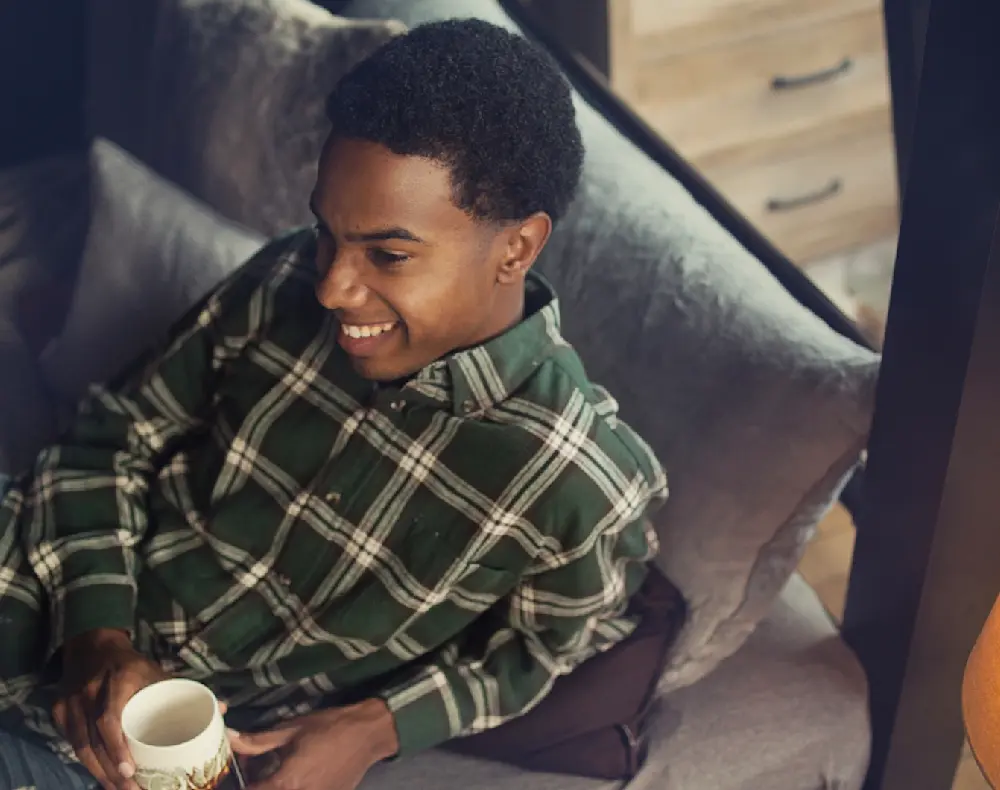 A man sits on the bottom bunk of an Adult Bunk Bed smiling and enjoying a cup of coffee.