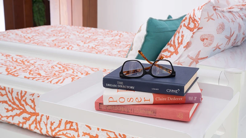 Close up of a white metal tray attached to the top bunk of a white Adult Bunk Bed with three books stacked on top of it and a pair of classes on the books.