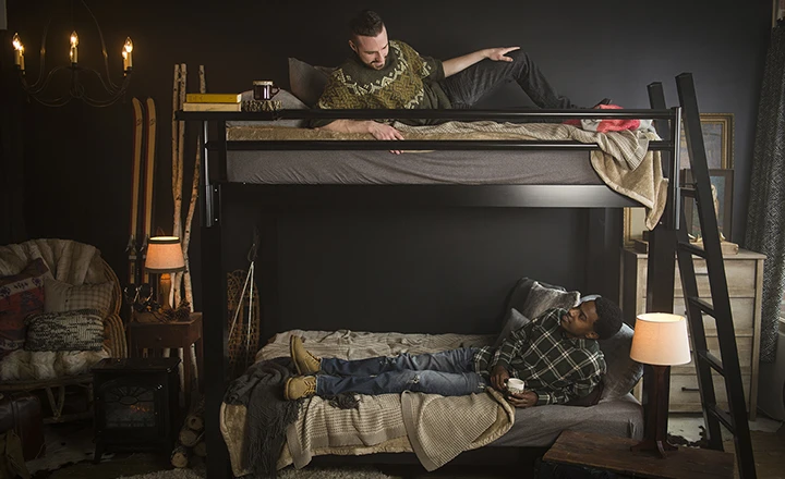 Two adult men on a black Adult Bunk Bed in a ski cabin bedroom. There is one on each bunk. They are looking up and down at one another and talking. They are seen directly from the left-hand side of the bed.