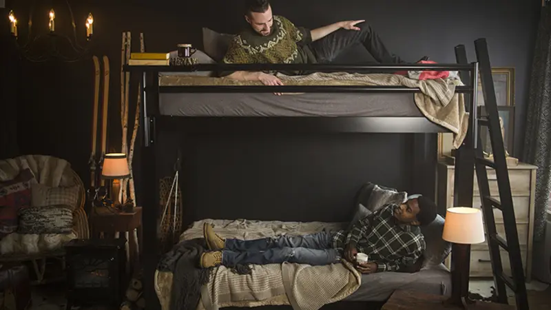 Two adult men on a black Adult Bunk Bed in a ski cabin bedroom. There is one on each bunk. They are looking up and down at one another and talking. They are seen directly from the left-hand side of the bed.