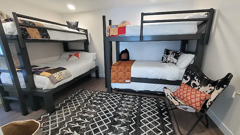 A neatly arranged bedroom featuring two black bunk beds, each with white bedding and colorful quilts. A patterned black and white rug covers the wooden floor, and a butterfly chair with a patterned cushion sits in one corner. The walls are white.
