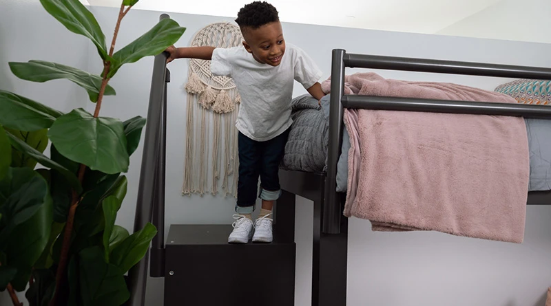 A young boy around 5-years-old standing on the top step of a four-step charcoal colored Wooden Staircase holding onto the Aluminum Handrail accessory and looking down at his mom.