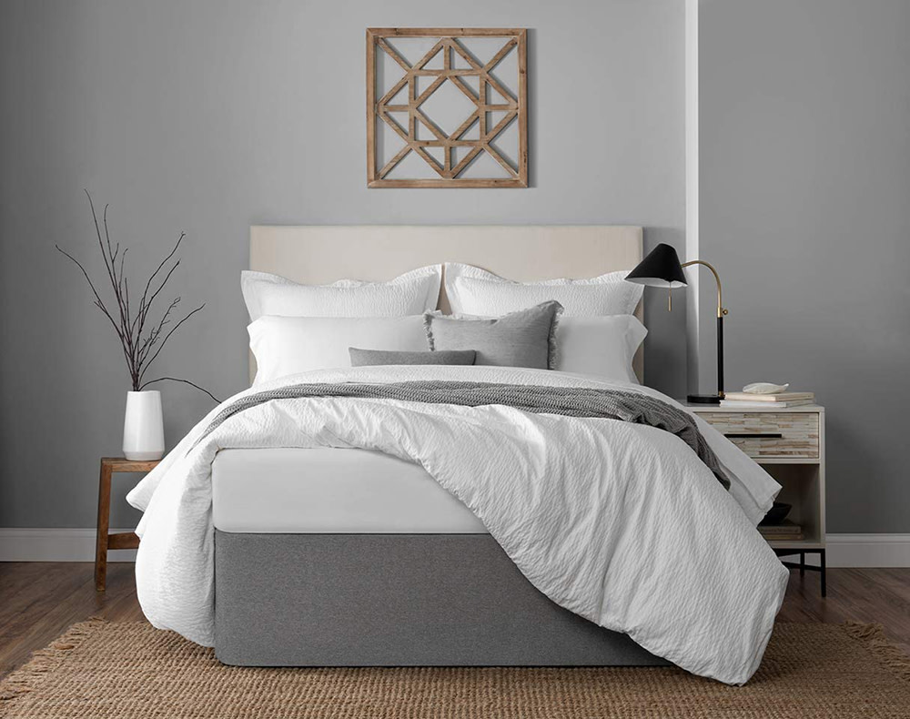 A queen size bed with a light beige fabric headboard and gray bed wrap around the bottom. Comfortable looking bedding in mostly white with some gray in a room with light gray walls.