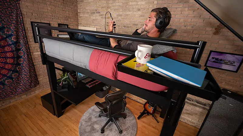 A large young man laying in his black queen size Adult Loft Bed listening to music on his phone.
