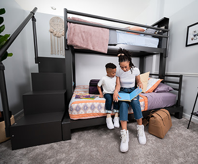 A charcoal Twin XL Over Queen Adult Bunk Bed with a young mother reading a book to her son on the bottom bunk.
