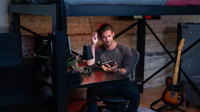 A man sits at a desk under a loft bed, holding a tablet and a stylus. He is surrounded by a lamp, a potted plant, and electronics. A guitar leans against a brick wall, and there are stairs leading up to the bed.