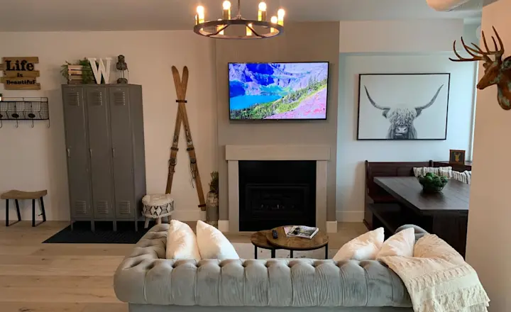 Photo of a living room in a condo in Whitefish, MT, facing the TV hanging above a fireplace from a view behind the gray couch. To the left, there is a small table nook with a high resolution large photo of a bison above it on the wall. To the right, a locker assembly with three tall lockers and two skis mounted to the wall.