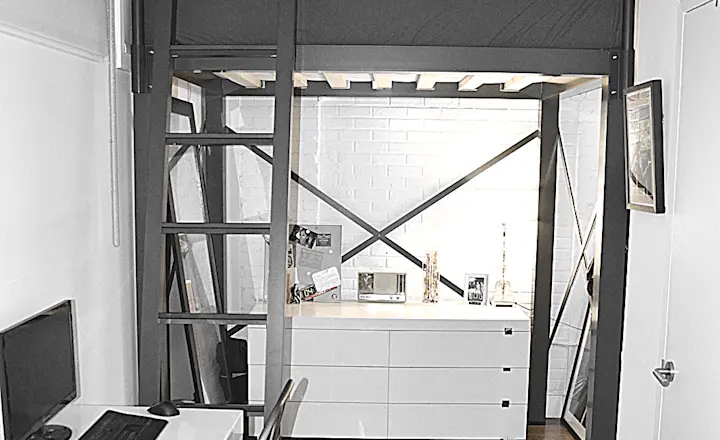 A modern, small apartment with a loft bed above a white dresser. There's a desk with a computer and chair in the foreground. The room has a white and gray color scheme, with wooden flooring.