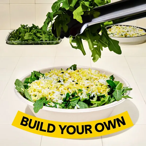 Tongs placing arugula into a CAVA bowl of arugula and saffron basmati rice. Text on the image says, "Build Your Own"