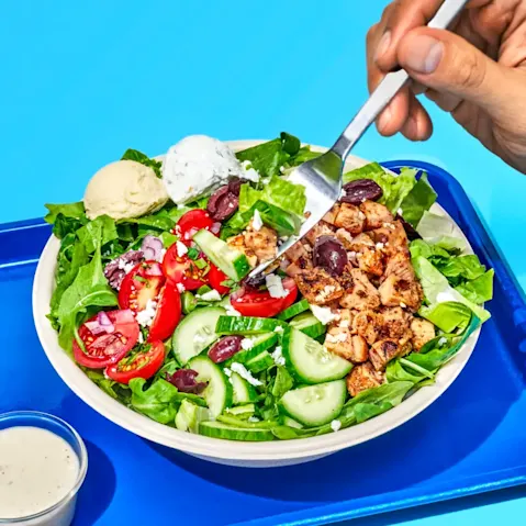 An image of a Greek Salad with someone's hand reaching for a bite with a fork