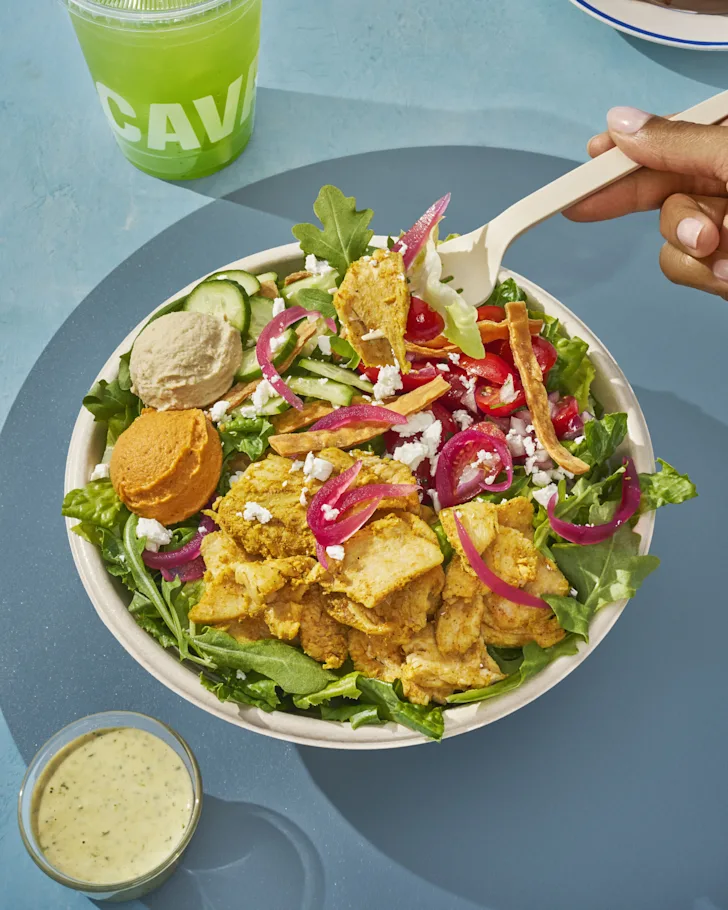 Garlicky Chicken Shawarma Bowl: A street cart-inspired chicken + rice bowl, bursting with bold ingredients. Chicken shawarma is paired with red pepper hummus, Crazy Feta®, corn, tomato + cucumber, pickled onions, fiery broccoli, arugula, saffron basmati rice, and the one-two, flavor-packed punch of skhug + garlic dressing.