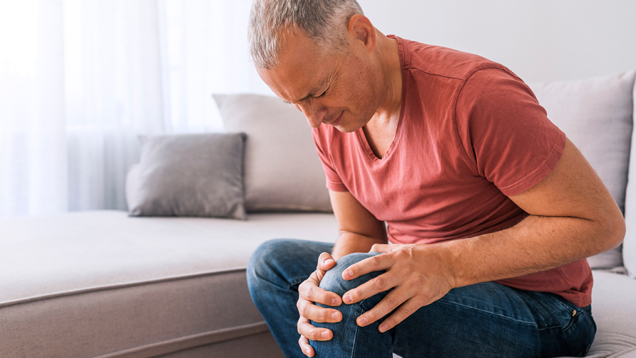 Mann sitzt auf der Couch und hält sein linkes Knie. Er leidet an Gelenkschmerzen.