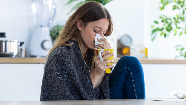 Frau mit Fieber und Schmerzen sitzt mit Tee und Taschentuch an einem Tisch.