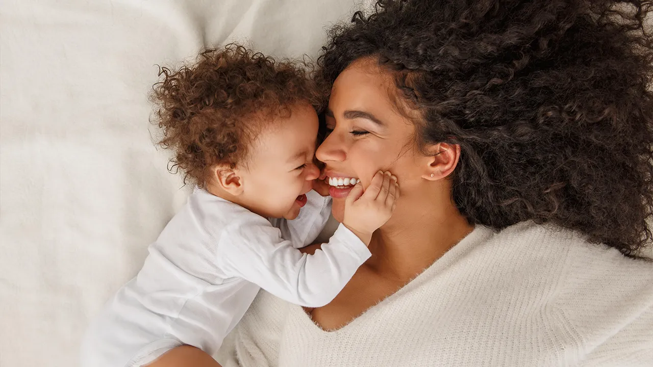 Junge Mutter liegt lachend mit ihrem Baby auf dem Bett.