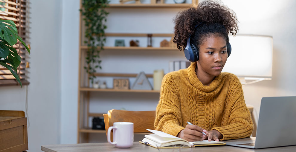 Young woman wearing headphones taking notes in a notepad while looking at a laptop screen.